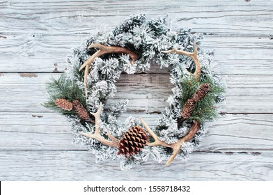 Flocked country or hunters Christmas wreath over white wooden background.  Made with pine cones, fir branches, and deer antlers and flocked with fake snow. Handmade decor for winter holidays. Top view - Powered by Shutterstock