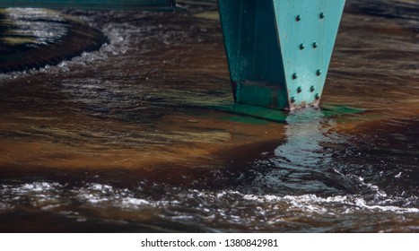 Flood water rolls over a concrete footing and around a rusty green column - Powered by Shutterstock