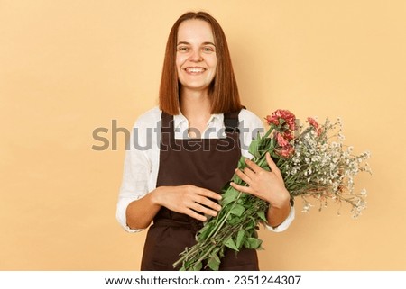 Floral designer. Plant store. Fresh blooms. Botanical beauty. Smiling young caucasian woman florist wearing brown apron holding flowers isolated over beige background