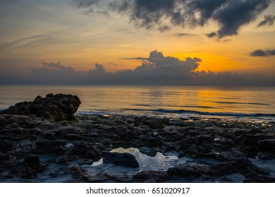 A Florida sunrise behind the clouds over a calm smooth ocean from a rocky beach with clouds reflecting in the tidal pools - Powered by Shutterstock