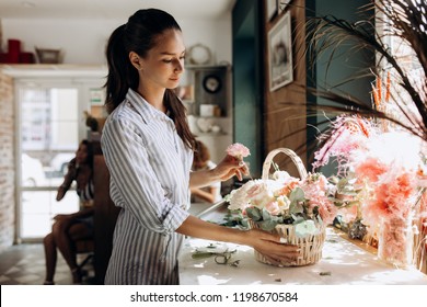 Florist dressed in a striped dress collections a bouquet in the basket from fresh flowers of pastel colors in the flower shop - Powered by Shutterstock