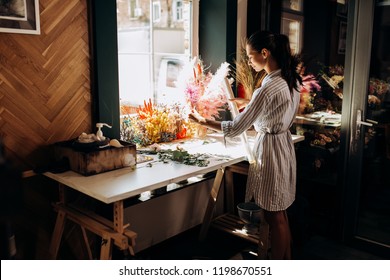 Florist dressed in a striped dress stands near the table with colorful dried flowers and holds transparent packaging envelope in the flower shop - Powered by Shutterstock