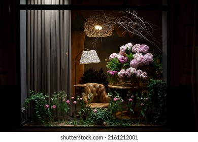 Florist Shop Window. Beautiful Decoration Of Luxurious Pink Flowers, Interior Items, Lighting. Showcase Window Of Floral Shop With Atmospheric Lights, Vintage Armchair. Dark Exquisite Stage Photo 