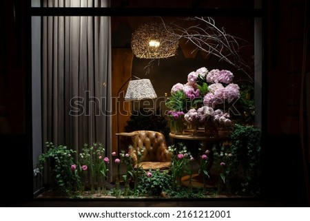 Florist shop window. Beautiful decoration of luxurious pink flowers, interior items, lighting. Showcase window of floral shop with atmospheric lights, vintage armchair. Dark exquisite stage photo 