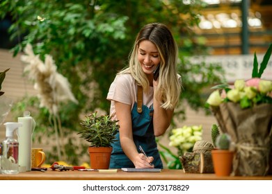 Florist talking with client on the smartphone and taking an order while standing next to the counter surrounded with flowers and plants in a flower shop - Powered by Shutterstock