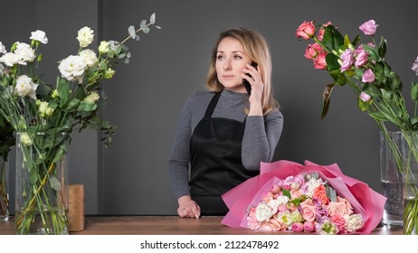 Florist Woman In Black Apron Informs Customer Via Mobile Phone About Arranged Flower Bouquet For Wedding On Wooden Table In Flowershop.