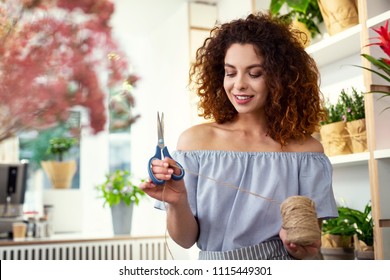 Floristic design. Nice pleasant woman cutting a piece of rope while working on a floristic design - Shutterstock ID 1115449301