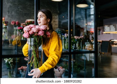 Floristics, business, decoration concept. Young inspiration woman in yellow sweater in grey apron holding vase with roses in modern floral shop dark interior. - Powered by Shutterstock