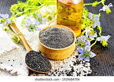 Flour Of Black Caraway In A Bowl, Seeds In A Spoon On Burlap, Oil In Bottle And Twigs Nigella Sativa With Blue Flowers And Leaves On Black Wooden Board Background