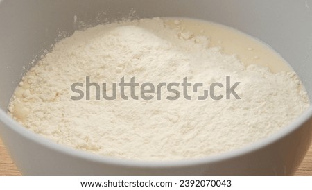 Flour pours into white bowl. A fine stream of white flour is added to the contents of a bowl, showcasing the preliminary steps in baking or cooking.