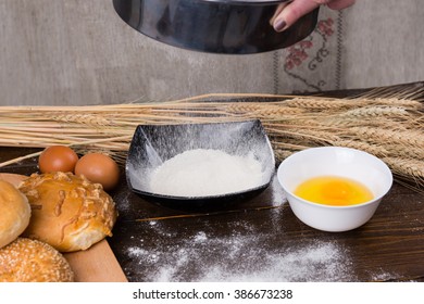 Flour sifter in action next to brown eggs, yolks and various rolls on wooden table with dried wheat stalks in background - Powered by Shutterstock