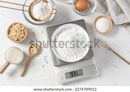 Flour in white bowl measuring on digital scale with cake or bakery ingredients and utensil on marble kitchen table, top view