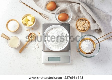 Flour in white bowl measuring on digital scale with baking ingredients and utensil on marble kitchen table, top view