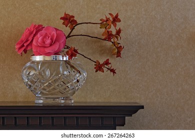 Flower arrangement, including camellia flowers, in crystal vase on carved wooden mantelpiece against plain wallpaper [landscape format].
