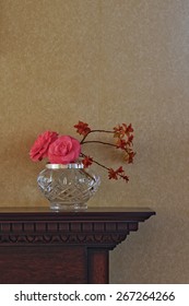 Flower arrangement, including camellia flowers, in crystal vase on carved wooden mantelpiece against plain wallpaper [portrait format].