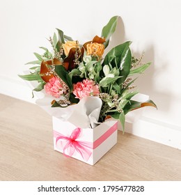 Flower Bouquet In White Box With Pink Ribbon. Wooden Flooring. White Wall Behind. Pink Flowers, Green Leaves. Side Angle.