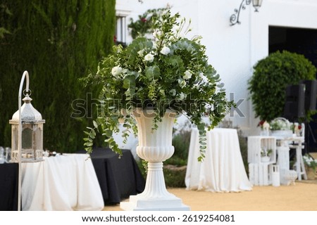 Flower pot with eucalyptus branches and white flowers in the decoration for an outdoor wedding. There are catering tables and lanterns in the decoration of the event