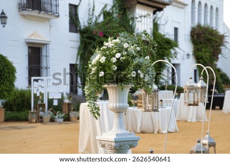 Flower pot with eucalyptus branches and white flowers in the decoration for an outdoor wedding. There are catering tables and lanterns in the decoration of the event