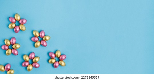 Flower shaped pile of Easter candy eggs wrapped in pink and golden foil on blue background. Happy Easter, springtime concept. Preparation for holiday. Simple minimalism. Top view, copy space