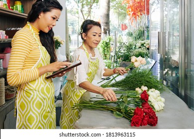 Flower Shop Owner Working On Tablet Computer When Florist Making Bouquet For Customer