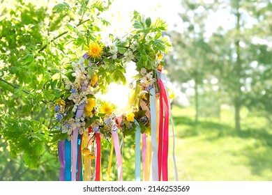 flower wreath with colorful ribbons on tree in garden, green natural background. floral decor, Symbol of Beltane, Wiccan Celtic Holiday beginning of summer season. pagan witch traditions, rituals.