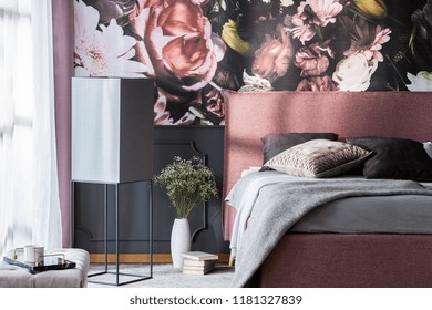 Flowers and books next to pink bed with grey pillows in patterned bedroom interior. Real photo