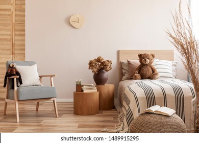 Flores de jarrón marrón sobre una mesa de noche de madera al lado de una cama individual con cama de matrimonio a rayas con osito de peluche