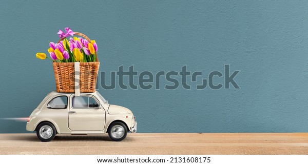 Flowers delivery. White Retro toy car carrying\
flowers tulips in a wicker basket. Valentine day, womens day. Place\
for text.