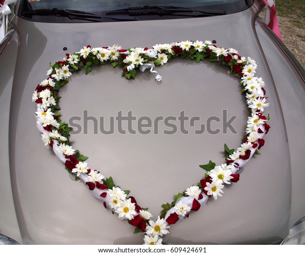 Flowers of different\
kinds are in the shape of a heart on the hood of the car. A couple\
is getting married.