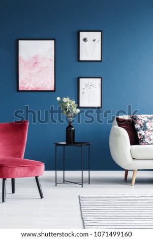 Flowers on black table between red chair and sofa in blue living room interior with posters