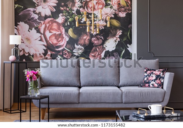 Flowers on black table and grey sofa in living room interior with lamp and feature wall wallpaper. Real photo