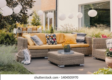 Flowers on rattan table near couch with patterned cushions on the terrace with lanterns. Real photo
