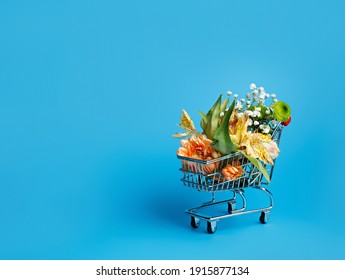 Flowers In Small Shopping Trolley On Blue Background. Flower Delivery Concept. Sale, Shopping For The Holidays Mothers Day, Women's Day, Valentines Day Or Birthday. Copy Space.