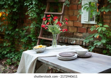 Flowers In A Vase At Home. Interior Terrace, Patio. Wooden Table And Plates For Garden Party Or Dinner. Table For Lunch Outside In Garden In Yard Of House. A Bouquet Of Red Poppies In A Glass Vase. 