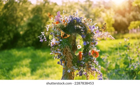 flowers wreath on wooden post and butterflies on sunny meadow natural background. Floral crown, symbol of Summer Solstice Day, Midsummer holiday. witch tradition, wiccan ritual. Litha sabbat.
