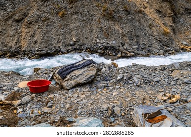 Flowing river with rapids cuts through rugged terrain in Kyrgyzstan. Red basin, dark clothing, and sandals on riverbank suggest human activity in nature.