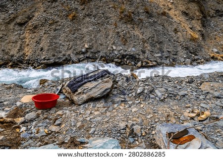 Flowing river with rapids cuts through rugged terrain in Kyrgyzstan. Red basin, dark clothing, and sandals on riverbank suggest human activity in nature.