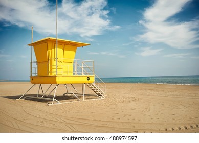 Flowy white clouds above a sunlit yellow lifeguard post on an empty sandy beach near quiet sea