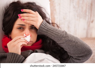 Flu. Closeup image of frustrated sick woman with red nose lying in bed in thick scarf holding tissue by her nose and touching her head - Shutterstock ID 245721847