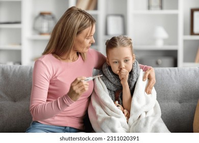 Flu Concept. Worried mother checking her daughter's temperature at home, caring mom holding thermometer and embracing sick little girl covered in blanket, female kid coughing into fist - Shutterstock ID 2311020391