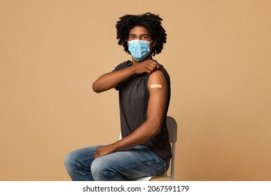 Flu Vaccination. Young Black Man In Protective Medical Mask Showing Arm With Plaster After Getting Coronavirus Vaccine Shot, Vaccinated African American Guy Posing Over Beige Background, Copy Space