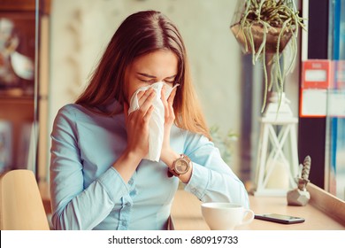 Flu. Young woman got nose allergy, flu sneezing nose sitting at the table in a trendy cafe coffee shop with a cup of hot beverage beside