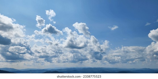 Fluffy clouds glide gracefully across the sky, casting playful shadows on the majestic mountains and lush green grasslands below on a sunny summer day. The play of light and shadow creates a mesmerizi