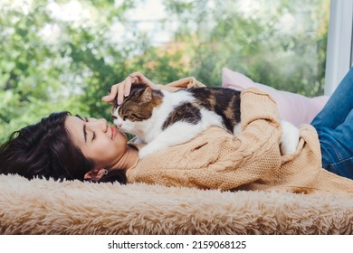 Fluffy and cute scotish cat laying down on asian woman chest while she lay dow on brown carpet at living room, touching cat with love feeling.