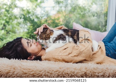 Fluffy and cute scotish cat laying down on asian woman chest while she lay dow on brown carpet at living room, touching cat with love feeling.