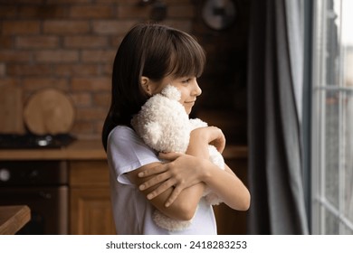 Fluffy friend. Smiling little girl stand by kitchen window alone hug plush teddy bear wait for mother parents coming back home. Adorable small child embrace favorite toy look at distance dream imagine - Powered by Shutterstock