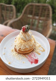Fluffy pancake stack topped with a mountain of grated cheese, a scoop of vanilla ice cream, and a fresh strawberry. Served on a white plate with caramel sauce and strawberry jam in an outdoor cafe.