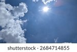 Fluffy white clouds frame the left side of a deep blue sky while the sun shines brightly with a starburst effect. A dramatic skyscape abstract background with large copy space.