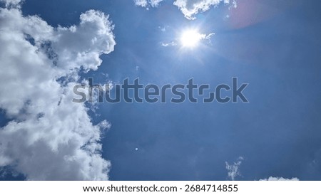Fluffy white clouds frame the left side of a deep blue sky while the sun shines brightly with a starburst effect. A dramatic skyscape abstract background with large copy space.