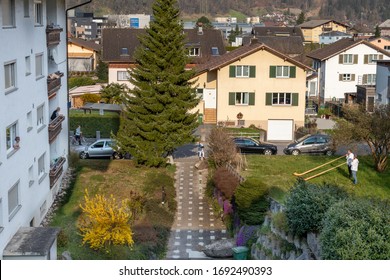 Flums, Switzerland - 03.04.2020: A Senior Is Given A Birthday Visit In The Form Of An Alphorn Duet While She Listens From The Balcony Of An Apartment Building In Flums.